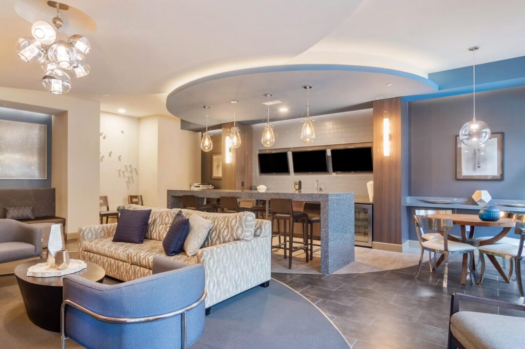 Interior clubhouse with couches and chair. Barstool seating and tvs mounted on the wall. Kitchen area with sink and mini fridges.
