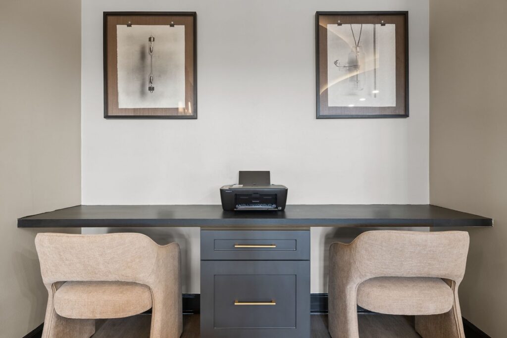A home office desk with a printer centered on the counter, two upholstered chairs underneath, and two framed abstract prints hanging on the wall above.