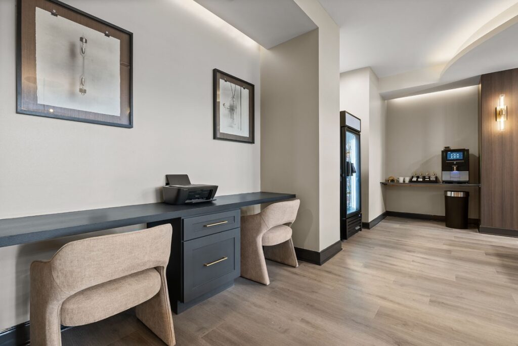 A hotel business center with a desk, two chairs, a printer, a small beverage refrigerator, and a coffee station.