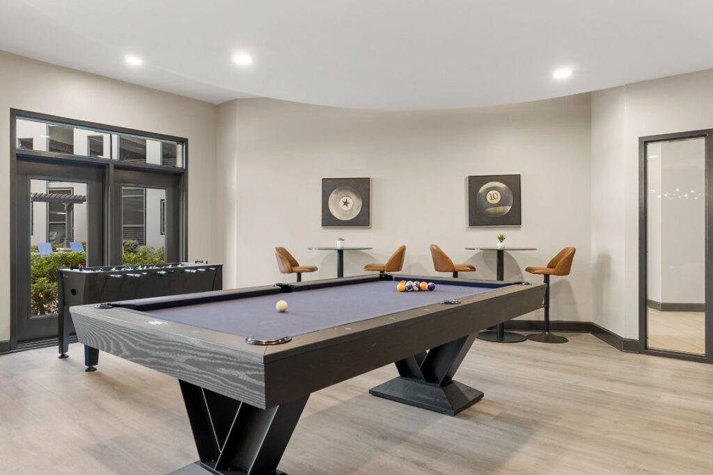 A game room featuring a black pool table, a foosball table, and two small cafe tables with tan bar stools.