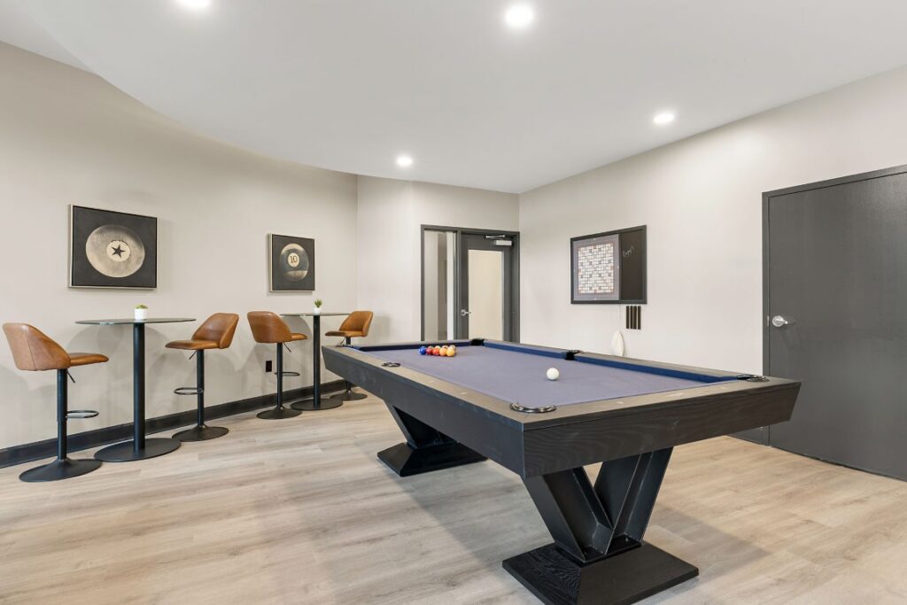 A game room featuring a blue pool table, two high-top bistro tables with bar stools, and a wall-mounted Scrabble board.