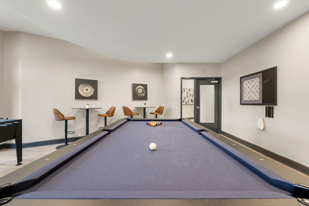 A game room with a blue felt pool table, two small bistro tables with bar stools, and a wall-mounted Scrabble board.