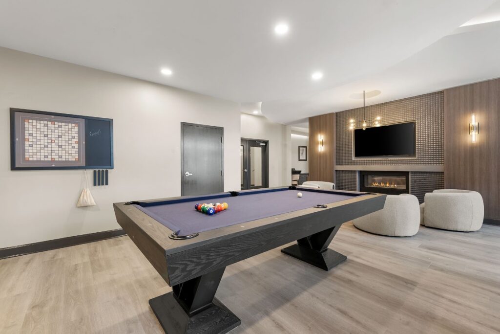 A pool table sits in a lounge area featuring a wall-mounted Scrabble board, a fireplace with a television above it, and two armchairs.
