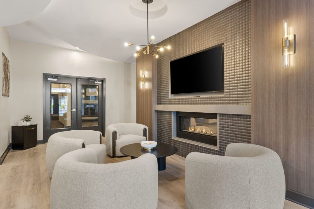 Four curved armchairs arranged around a circular coffee table in a lounge area featuring a fireplace, a wall-mounted television, and modern light fixtures.