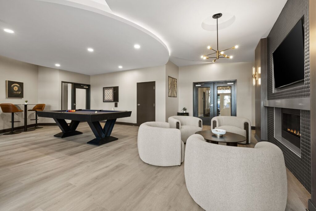 A lounge area featuring a pool table, four curved armchairs surrounding a small coffee table, and a wall-mounted television above a fireplace.