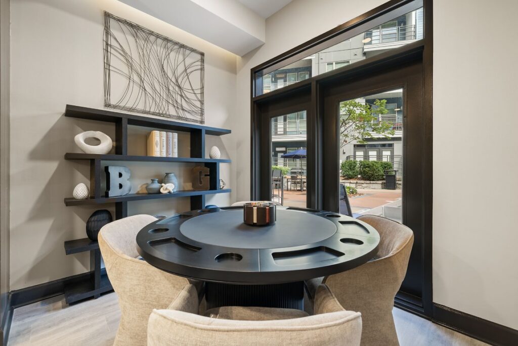 A circular black game table with four upholstered chairs in a room with a black wall shelf and a glass door looking out onto a patio.