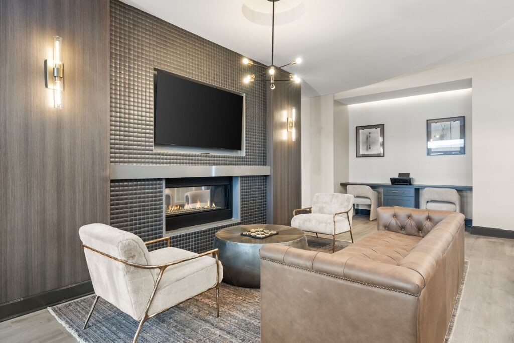 A lounge area with two cream-colored chairs, a tufted brown leather sofa, and a central coffee table facing a wall-mounted television above a fireplace.