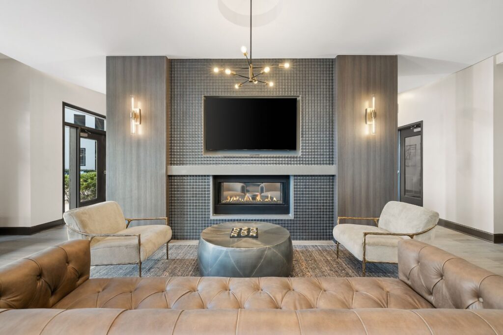 A lounge area with a central fireplace and wall-mounted television, flanked by two upholstered chairs, a round metal coffee table, and a leather sofa in the foreground.