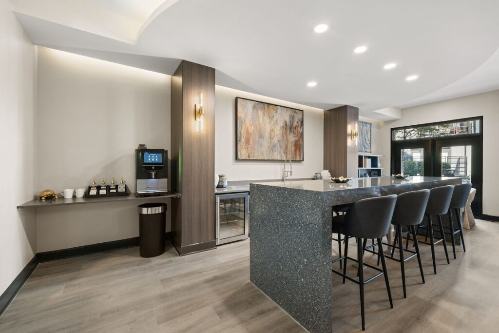 A modern kitchen or common area features a large, dark speckled island with four stools, a coffee station on a side counter, and abstract wall art.