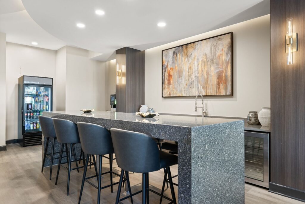 A kitchen island with a grey speckled countertop and four dark stools, featuring a small refrigerator, coffee machine, and large abstract painting on the wall.