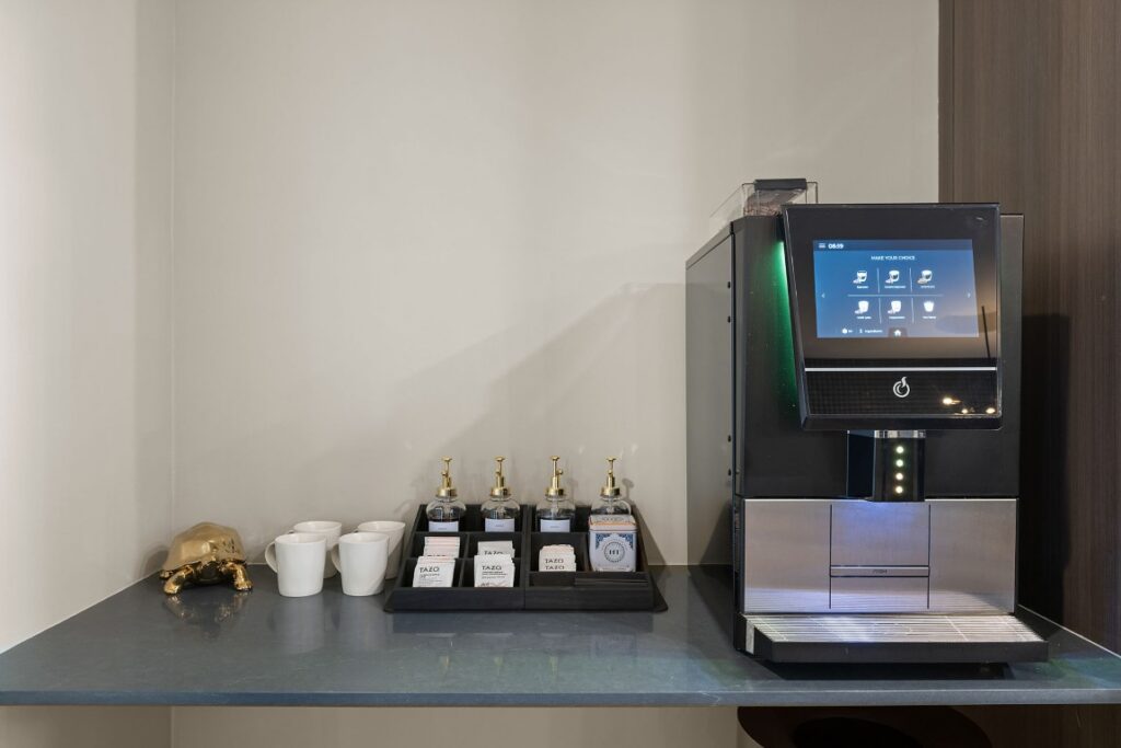 A touchscreen coffee machine next to a tray of tea bags, syrup bottles, and three white mugs on a counter.