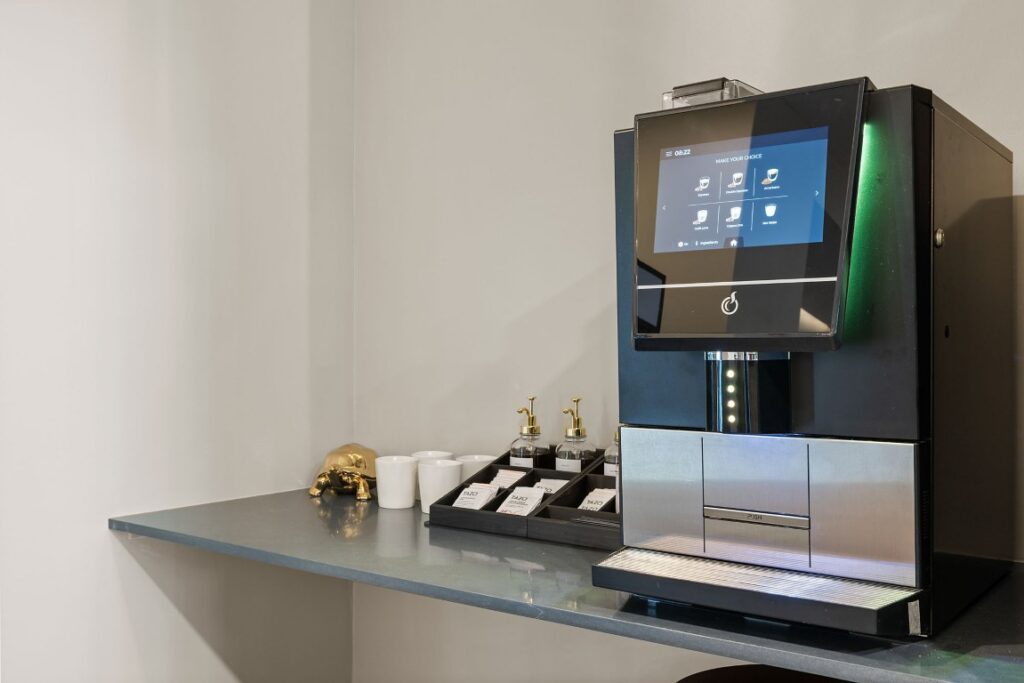 An automated coffee machine on a counter with white cups, syrup bottles, and a small decorative turtle.