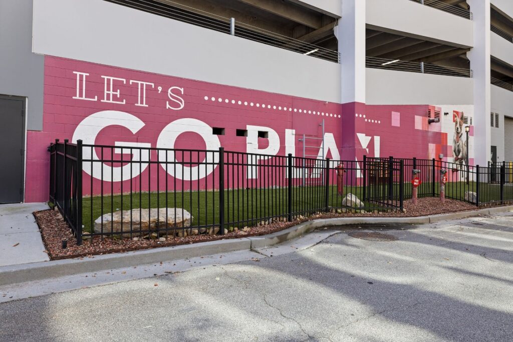 A fenced-in dog park area featuring a mural on a pink wall that reads LETS GO PLAY with a painted dog on the right side.
