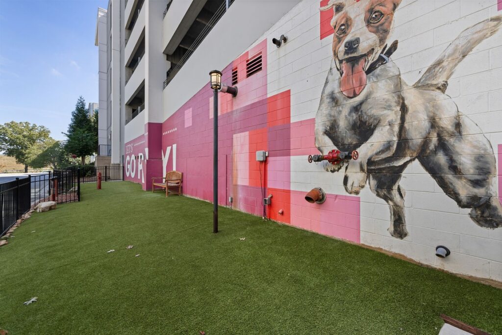A mural of a happy dog jumping on a pink and white geometric wall, located next to a small patch of artificial grass with a bench.