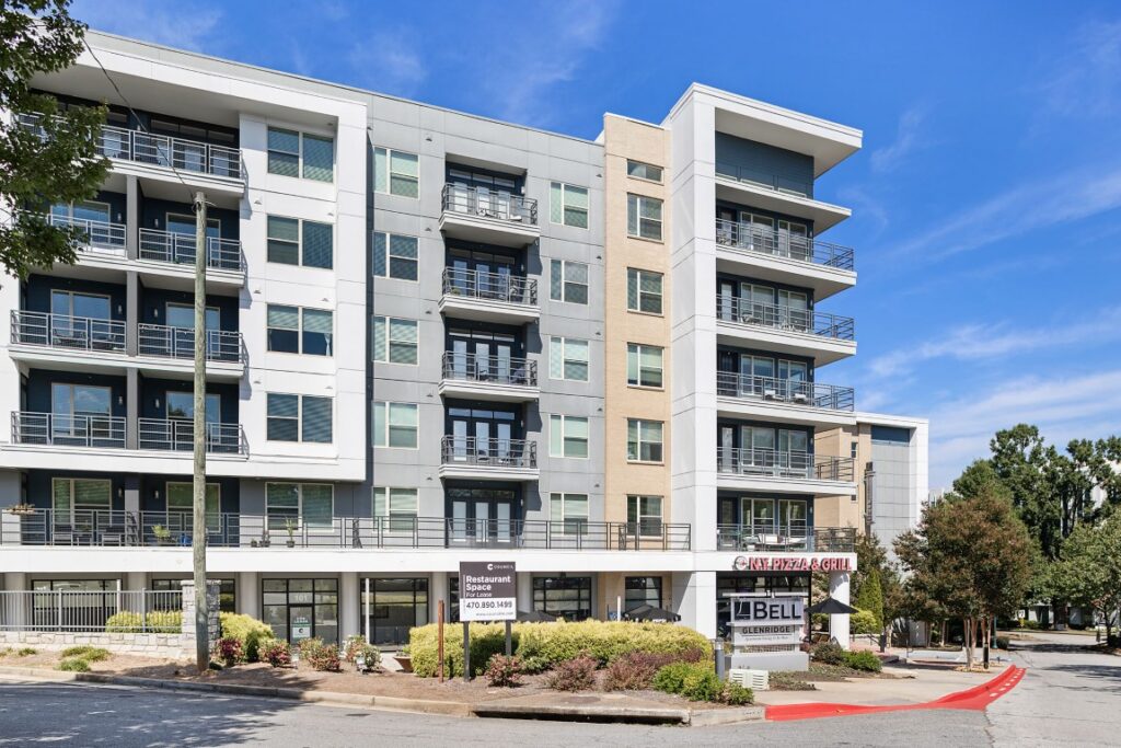 A multi-story apartment building with a street-level commercial area featuring a restaurant sign for NY Pizza & Grill and a leasing sign for restaurant space.