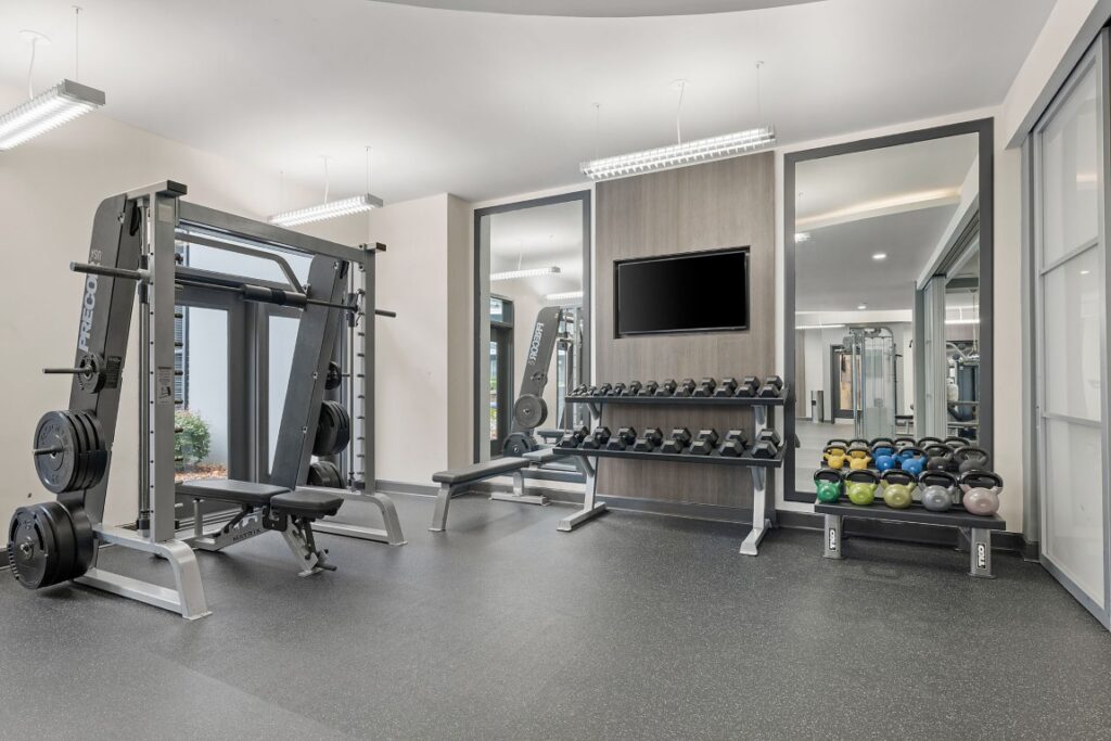 A fitness center with a weight machine, a rack of dumbbells, a row of kettlebells, and a wall-mounted television.
