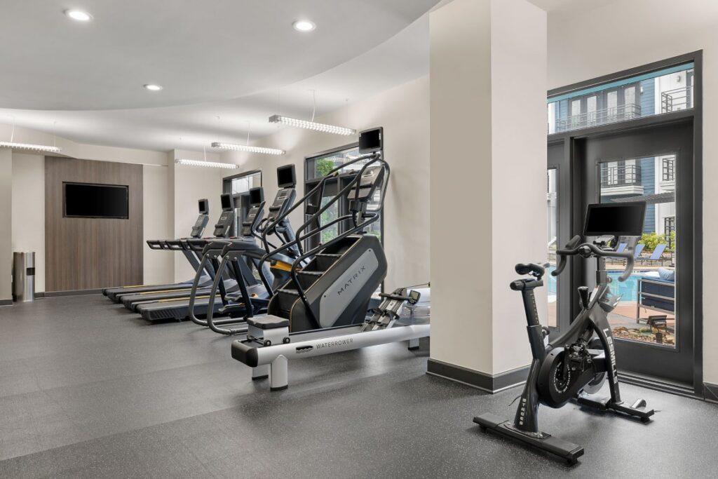 A gym interior featuring rows of treadmills, a stair climber, a rowing machine, and an indoor cycling bike near a patio door.