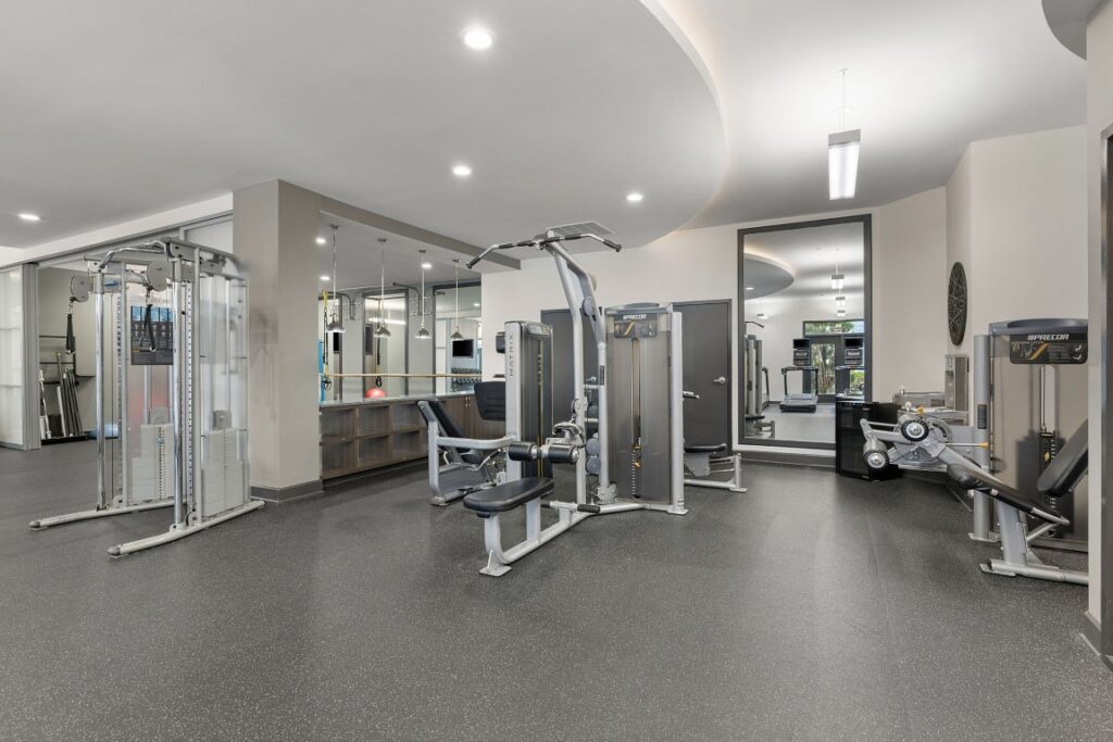 A gym interior featuring various weight training machines, including cable stations and a seated chest press, with a large mirror along the back wall.