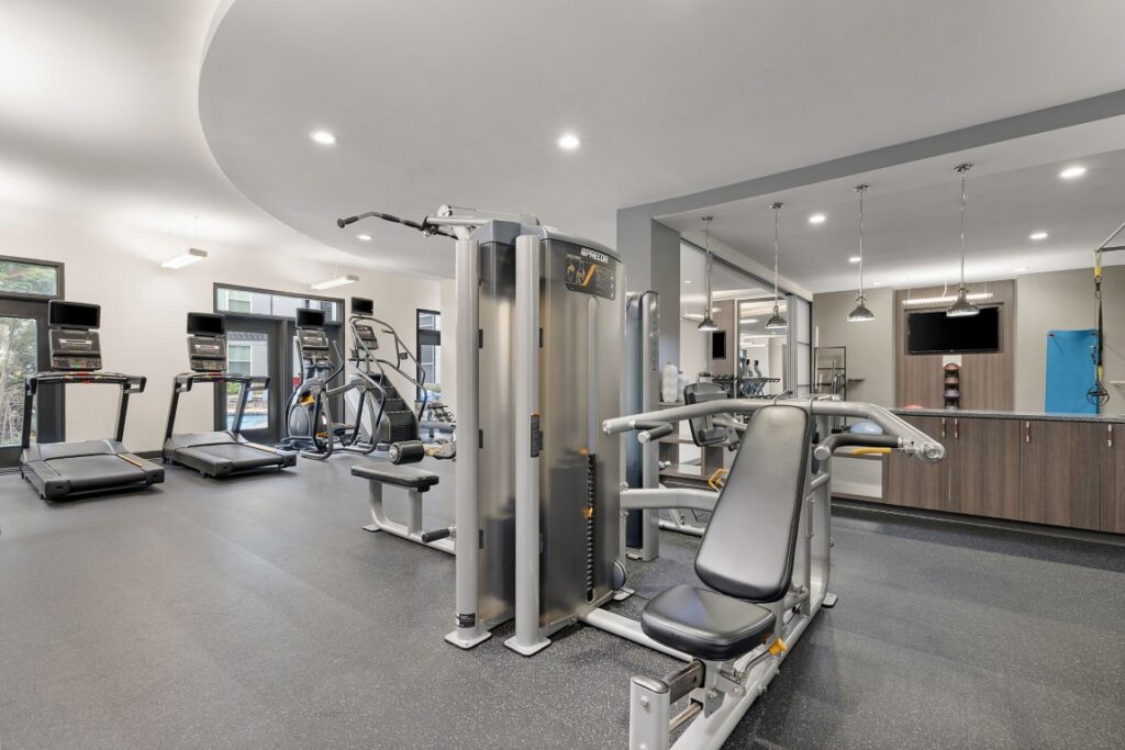 A gym facility featuring a multi-station weight machine in the foreground, with several treadmills and elliptical machines lined up against the windows.