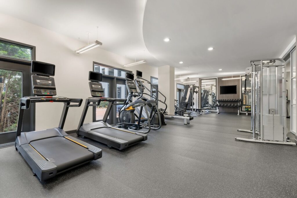 A gym featuring several treadmills, elliptical machines, and a cable strength training station on a dark rubber floor.