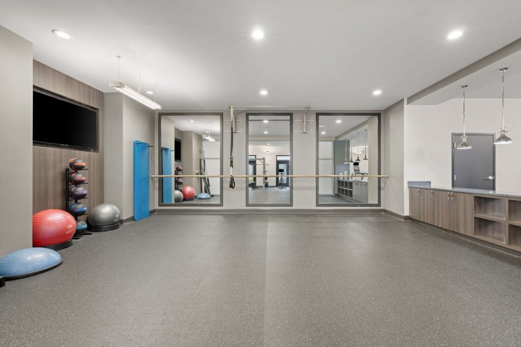 A gym studio featuring rubber flooring, a ballet barre attached to wall mirrors, a television, medicine balls, and a storage cabinet.