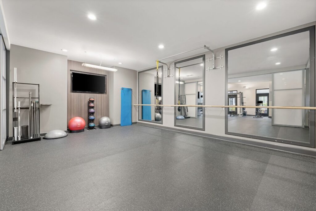 A gym studio featuring rubber flooring, a wall-mounted television, a rack of medicine balls, a ballet barre, and large wall mirrors.