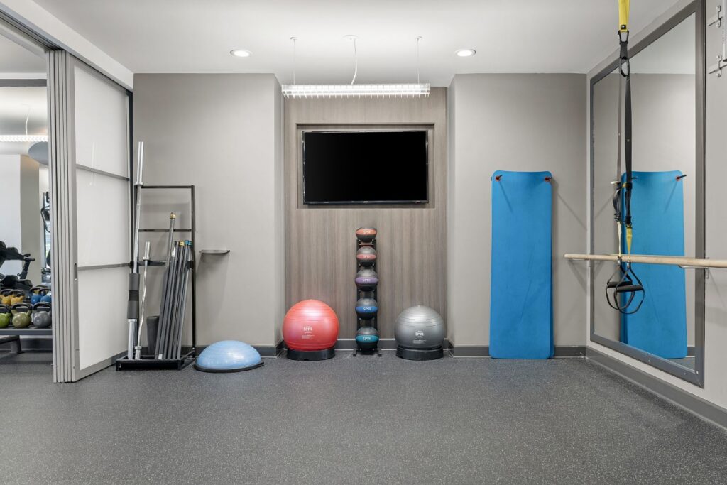Home gym with a wall-mounted television, a rack of medicine balls, yoga mats, resistance bands, a bosu ball, and a weight rack.