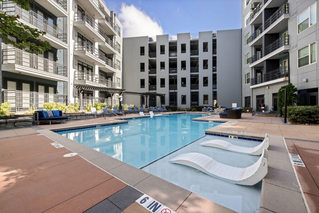 Outdoor swimming pool at an apartment complex, featuring two submerged lounge chairs in the shallow end and surrounding patio seating.