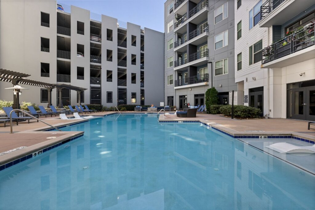 A courtyard swimming pool surrounded by lounge chairs, a fire pit, and multi-story apartment buildings.
