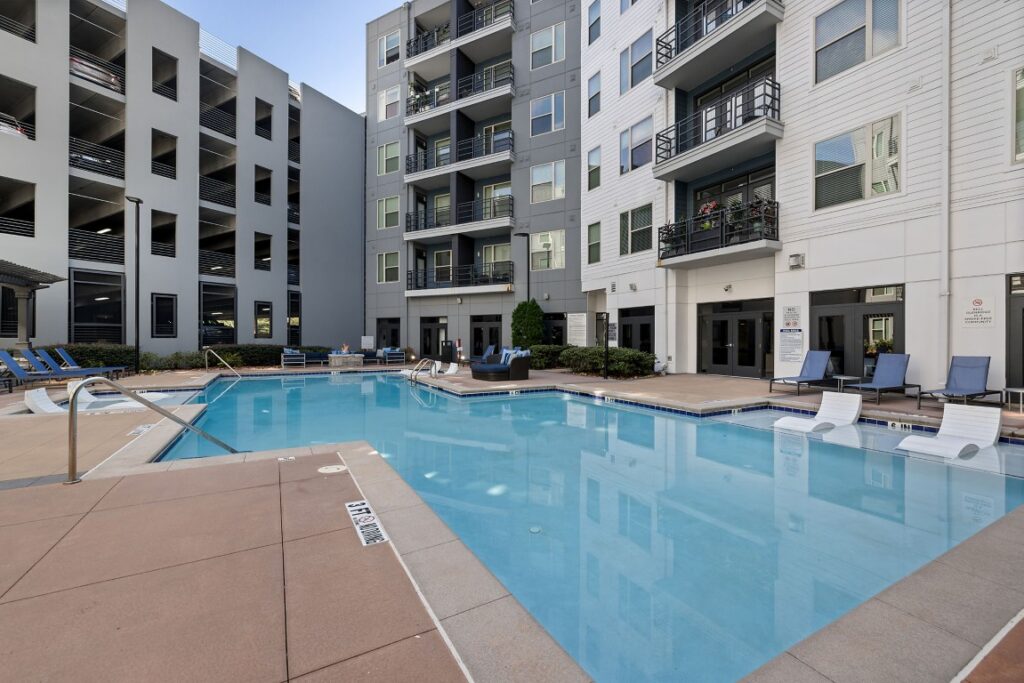 A swimming pool area at an apartment complex with lounge chairs on the deck and in the water, surrounded by multi-story residential buildings and a parking structure.