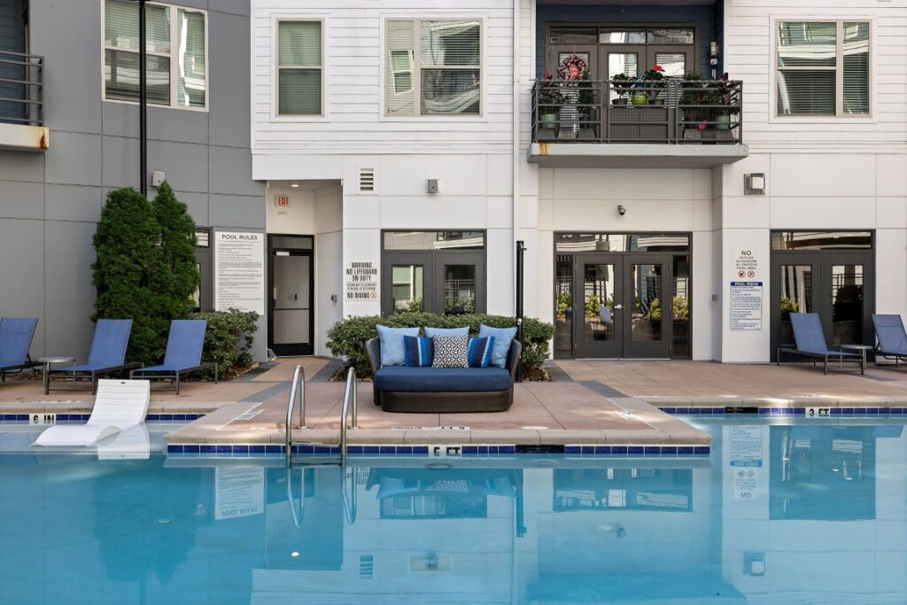 A swimming pool with lounge chairs and a round outdoor daybed on the deck, set against the side of a modern apartment building.