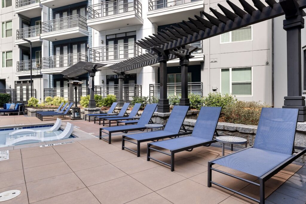 A row of blue lounge chairs on a pool deck in front of a modern apartment building with a pergola.