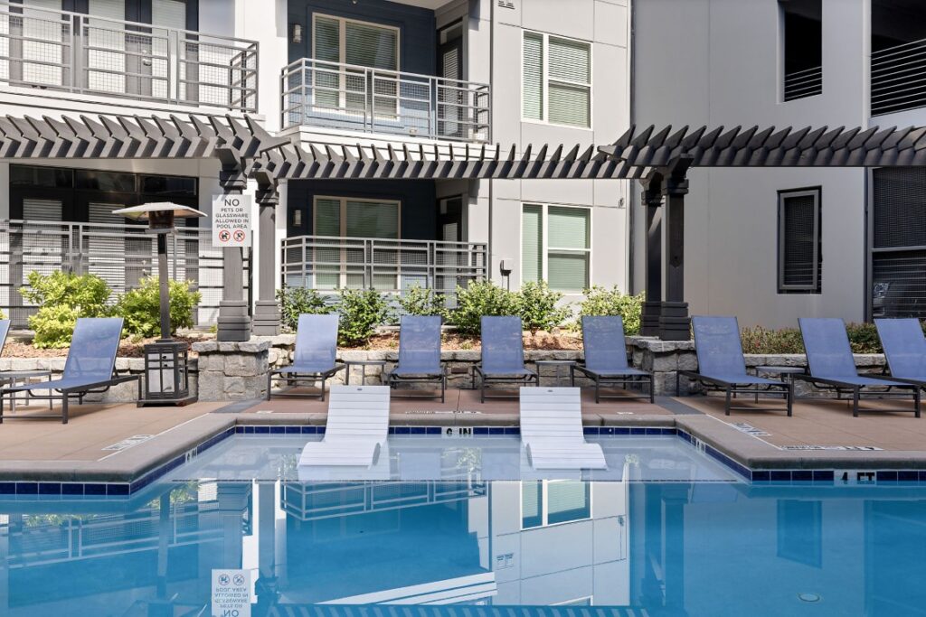 A swimming pool with two white submerged lounge chairs and a row of blue lounge chairs on the deck under a wooden pergola.