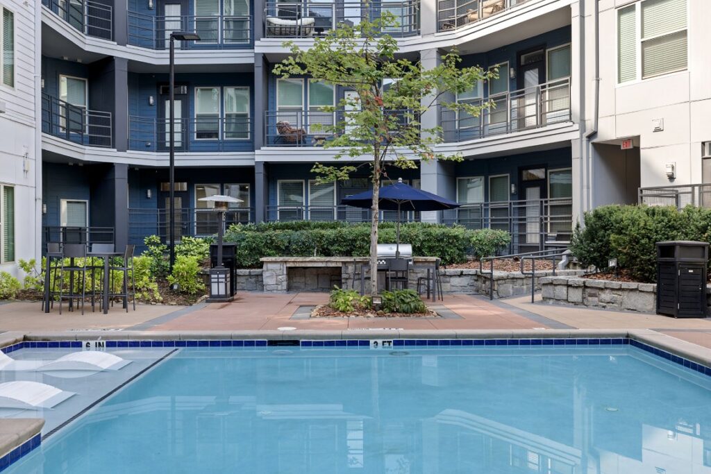 A swimming pool in a courtyard surrounded by a multi-story apartment building with balconies, outdoor seating, and a grill station.