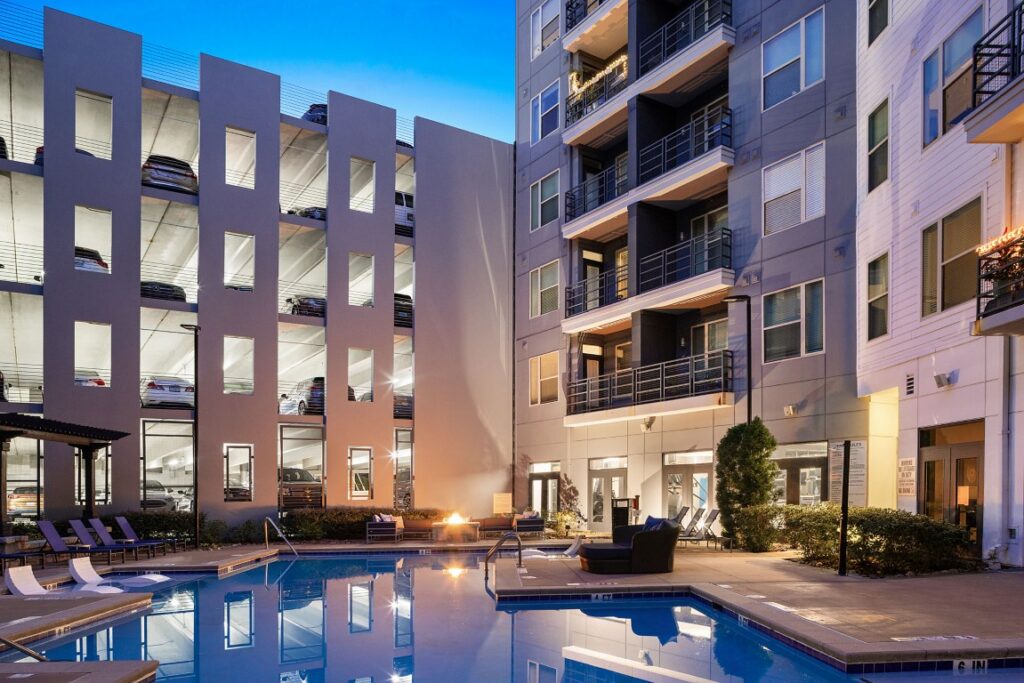 Swimming pool at an apartment complex at dusk, with an adjacent multi-level parking garage and balcony units.