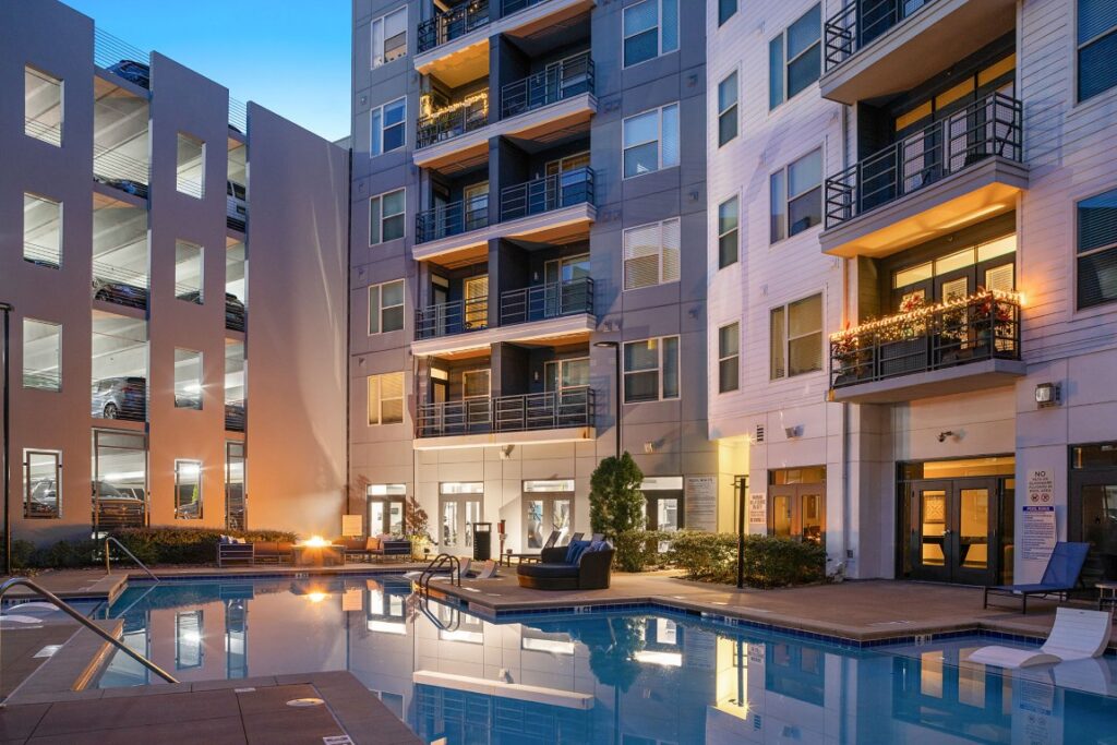 A swimming pool in a courtyard surrounded by modern apartment buildings and a multi-level parking garage at dusk.
