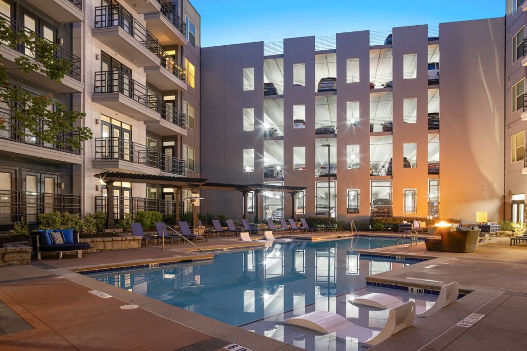 An apartment complex swimming pool at dusk, with lounge chairs on the deck and a multi-level parking garage in the background.