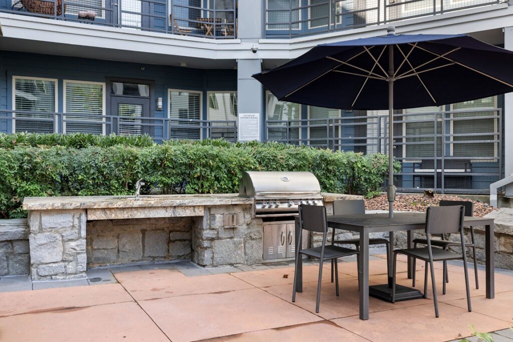 An outdoor kitchen area with a stone grill station, a dark blue patio umbrella, and a table with four chairs on a paved patio.