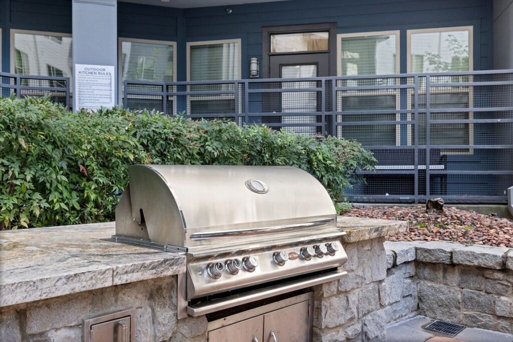 A stainless steel outdoor grill built into a stone counter, situated on a patio with a metal railing and bushes in the background.