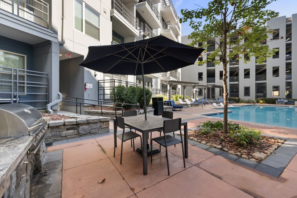 An outdoor patio with a dining table and black umbrella next to a swimming pool and a multi-story apartment building.