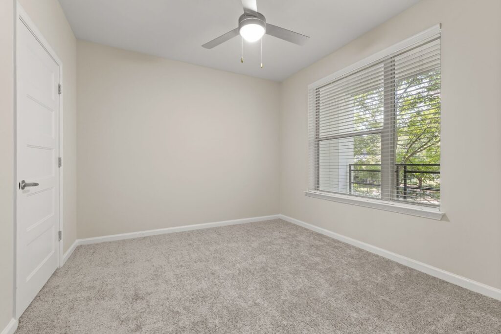 Empty room with beige walls, gray carpet, a ceiling fan, and a window with blinds looking out onto trees.