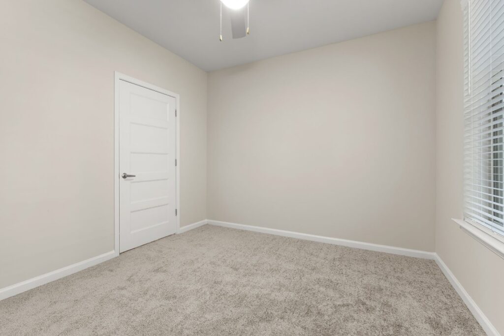 An empty room with beige walls, gray carpeting, a white door on the left, and a window with blinds on the right.
