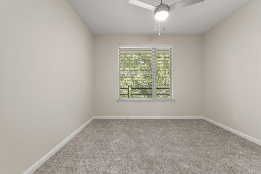 An empty room with beige walls, gray carpet, a ceiling fan, and a window with blinds looking out at trees.