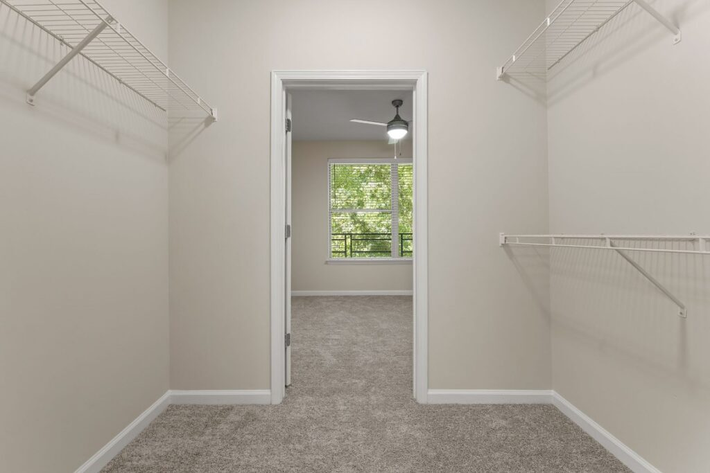A walk-in closet with wire shelving looking out into a bedroom with a carpeted floor, ceiling fan, and window.
