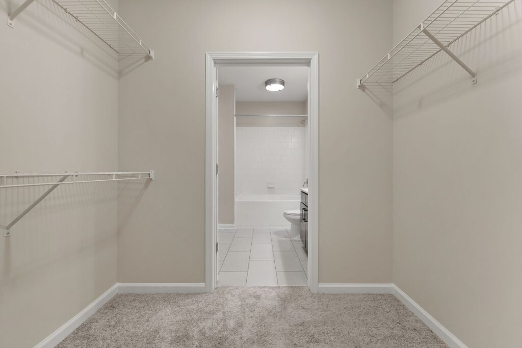 A walk-in closet with wire shelving looking through a doorway into a bathroom with a tub and toilet.