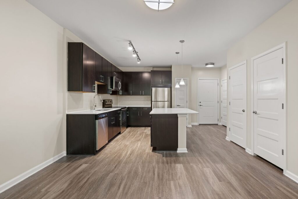 A kitchen with dark cabinets, stainless steel appliances, and a center island, opening into a hallway with multiple white doors.