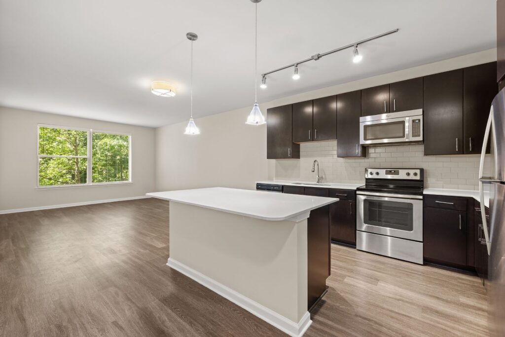 A kitchen with dark wood cabinets, a white island, and stainless steel appliances, opening to an adjacent living area with a window.