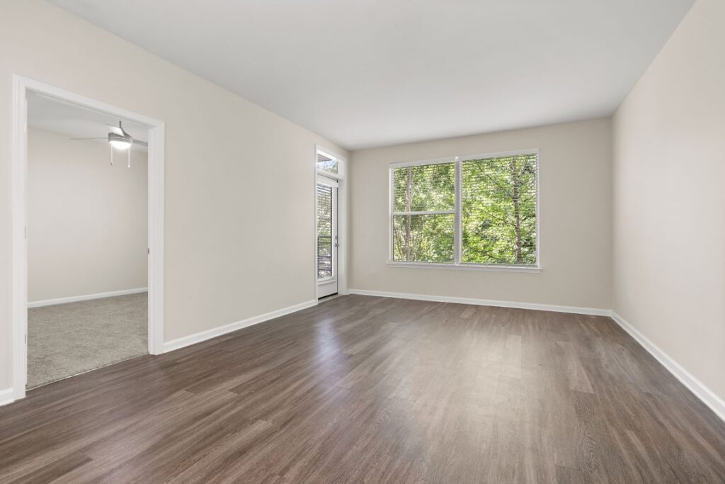 An empty room with wood-look flooring, beige walls, a window overlooking trees, and a doorway leading to a carpeted room with a ceiling fan.