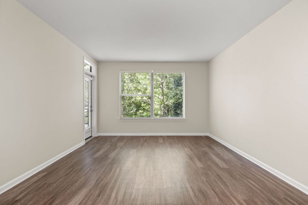 An empty room with beige walls, wood-look flooring, a large window with blinds, and a glass-paneled door leading outside.