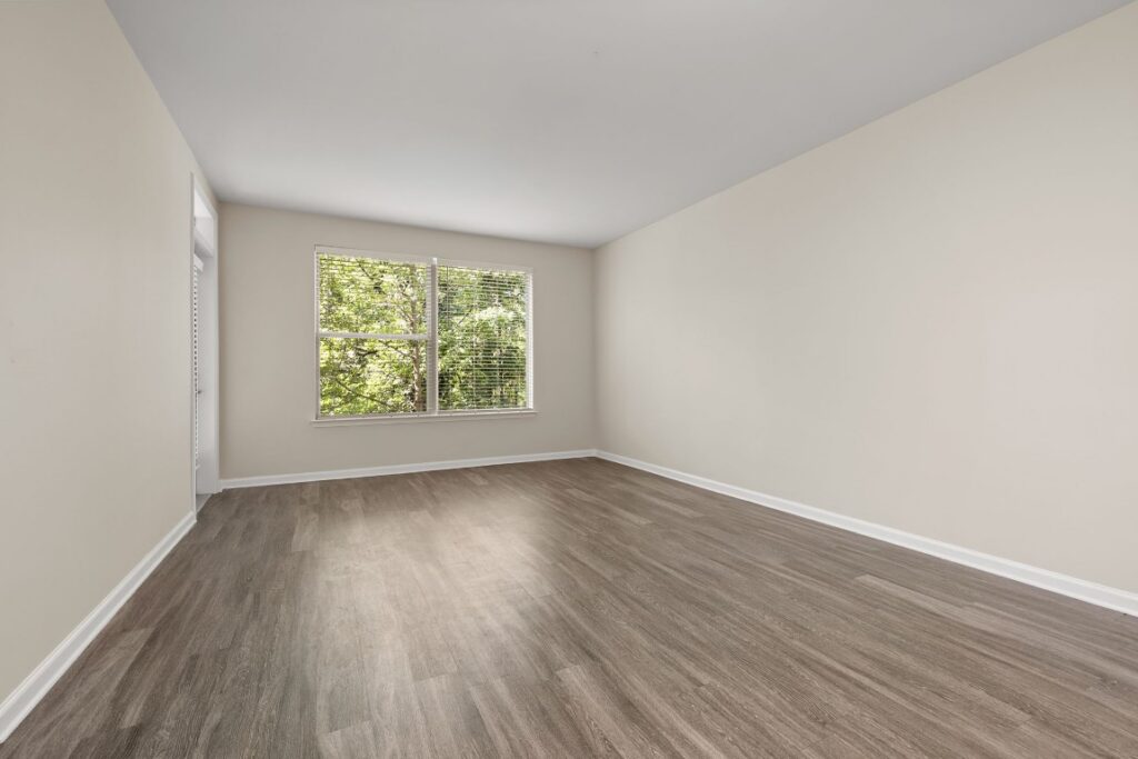 An empty room with light beige walls, wood-look flooring, and a large window looking out onto green trees.